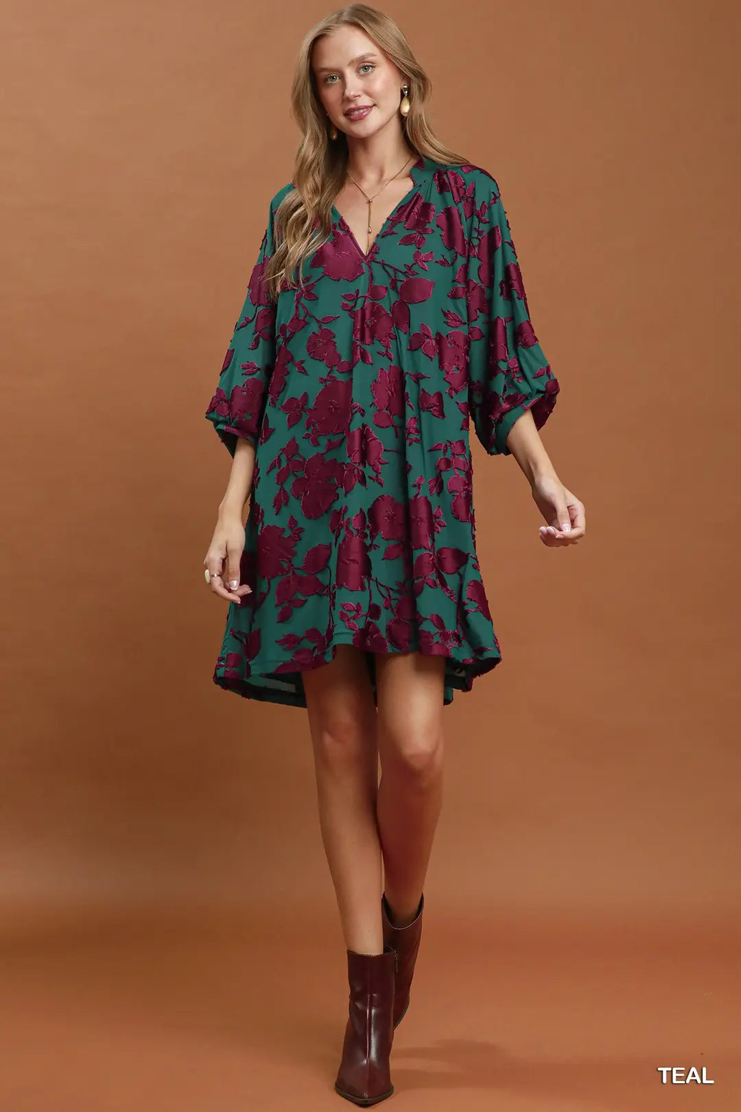 Woman wearing a teal and purple floral dress with brown boots on a brown background