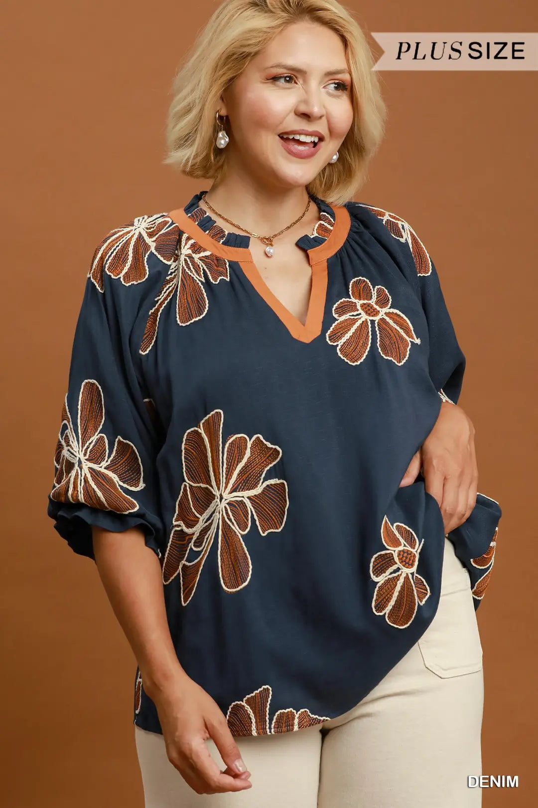 A woman wearing a navy blue blouse with a brown and white floral pattern and a tan V-neck trim, paired with cream pants, set against a solid brown background.