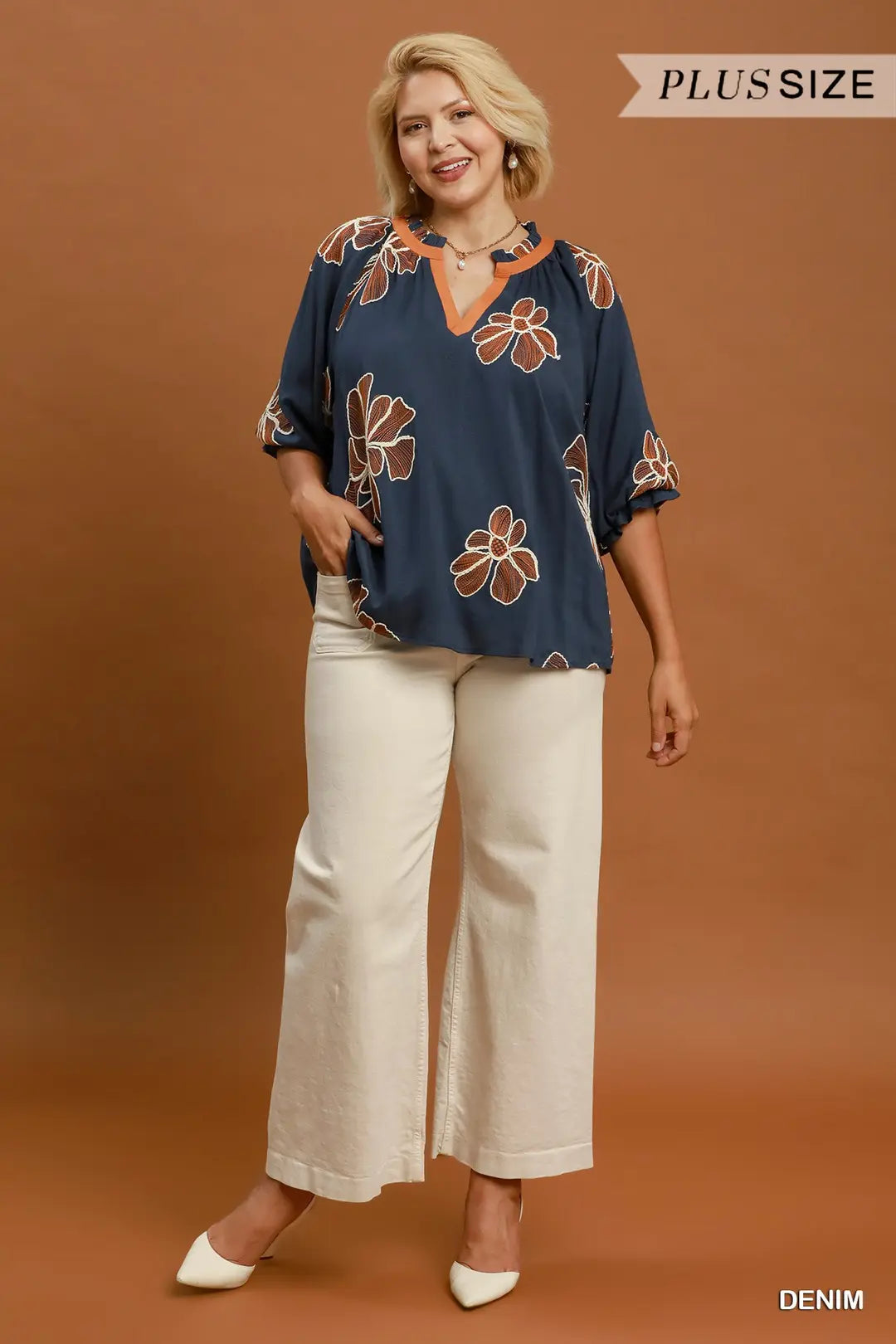 A woman modeling a navy blue plus-size blouse with a brown and beige floral pattern, paired with cream wide-leg pants, against a solid brown studio background. Text overlays read 'PLUS SIZE' and 'DENIM'.