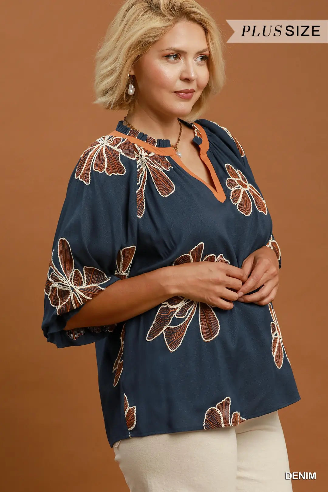 A woman wearing a navy blue plus-size blouse with brown and white floral patterns, paired with cream pants, against a solid brown studio background. A 'PLUS SIZE' banner is visible in the top right corner.