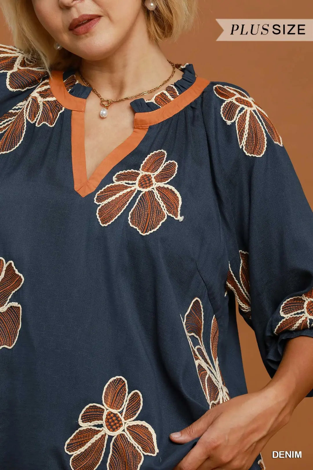A close-up of a woman wearing a navy blue plus-size tunic top with brown and beige floral embroidery and an orange split neckline. The image includes 'PLUS SIZE' and 'DENIM' text overlays.