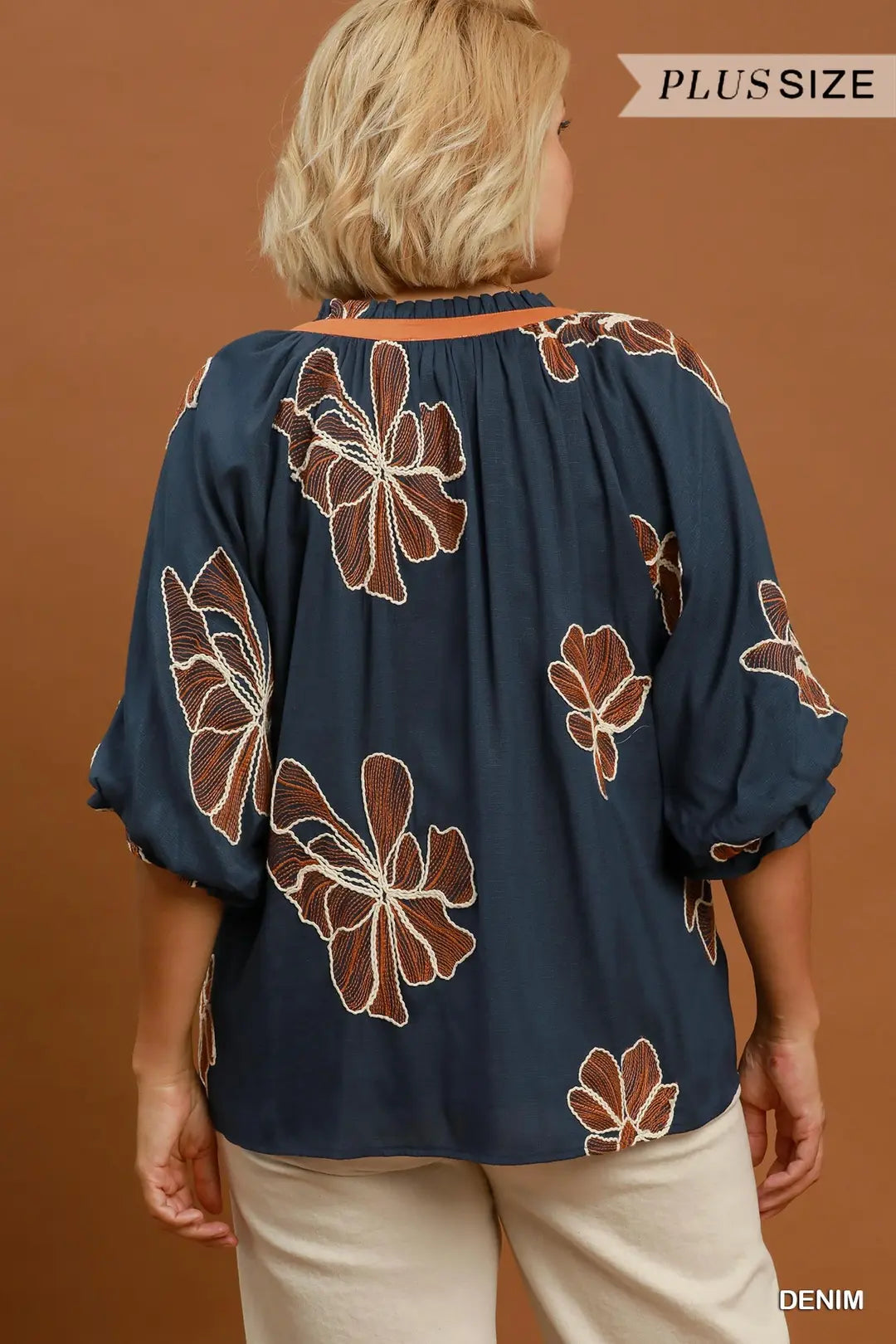 A woman viewed from the back wearing a navy blue plus-size blouse with a brown and white floral pattern, paired with light beige pants, against a solid brown background.