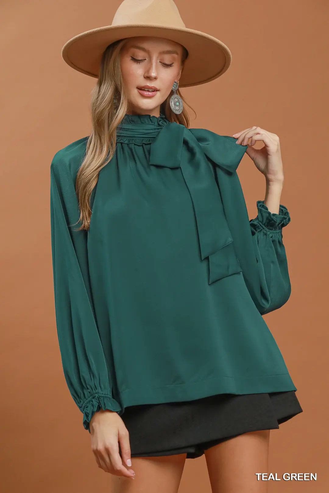A woman wearing a teal green long-sleeve blouse with a high ruffled collar and gathered cuffs, paired with a tan wide-brim hat and black shorts, against a solid warm brown background.