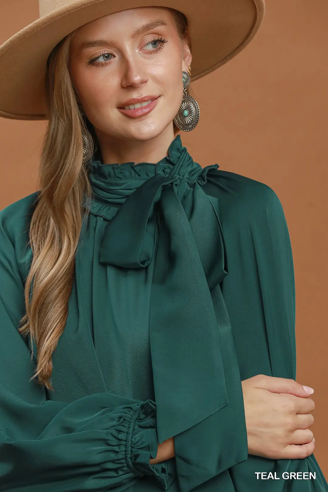 A woman wearing a teal green long-sleeve blouse with a ruffled high neck and large bow detail, styled with a wide-brim tan hat against a warm brown background.