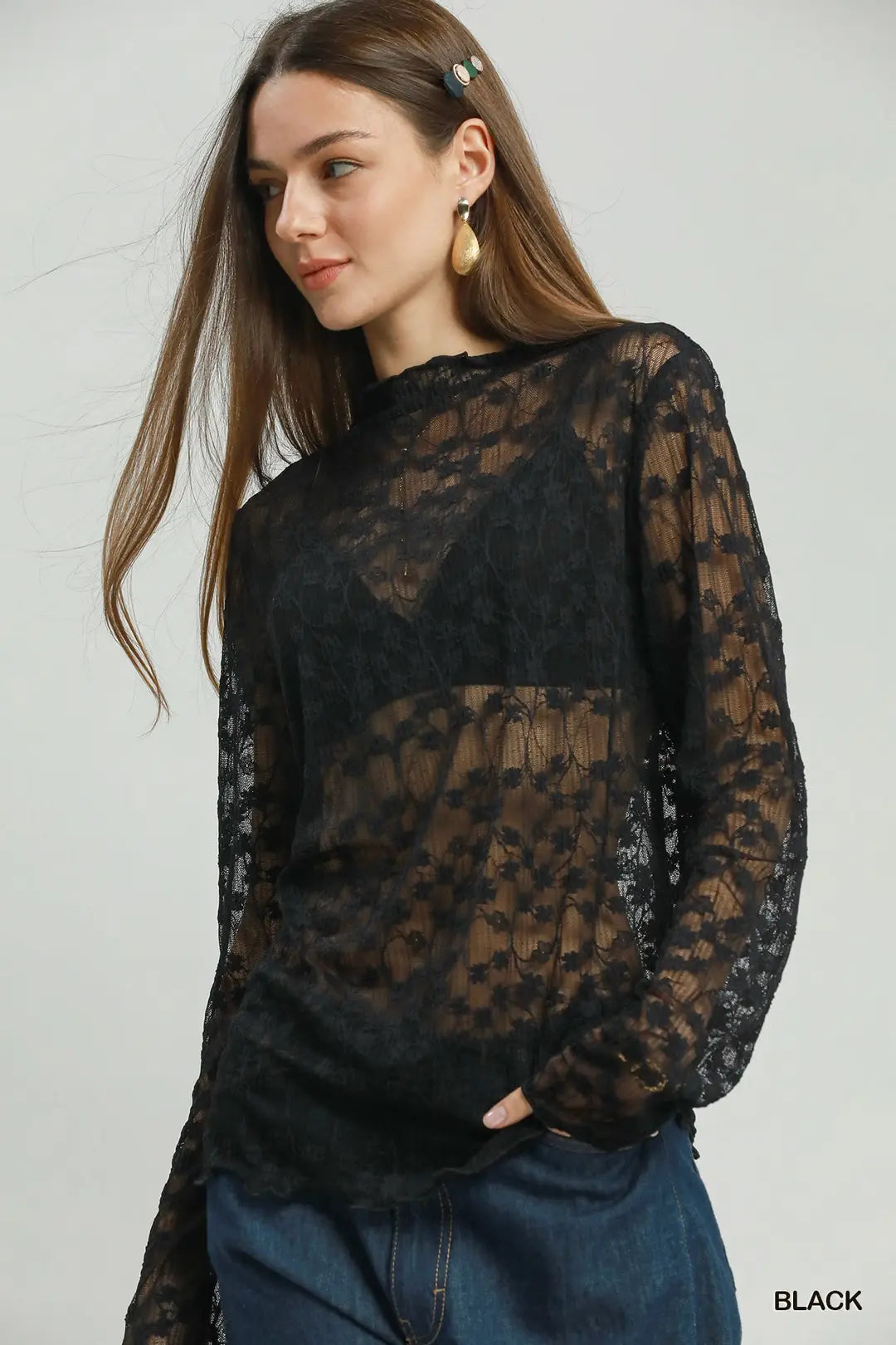 A woman wearing a sheer black lace long-sleeve top over a brown sheer underlayer, paired with blue jeans, against a light gray studio background.