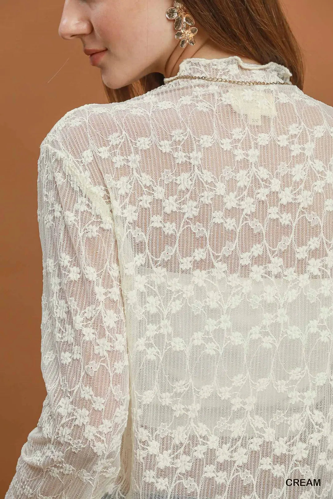 A close-up back view of a woman wearing a cream lace blouse with a ruffled high neckline and sheer floral pattern, against a warm brown background.