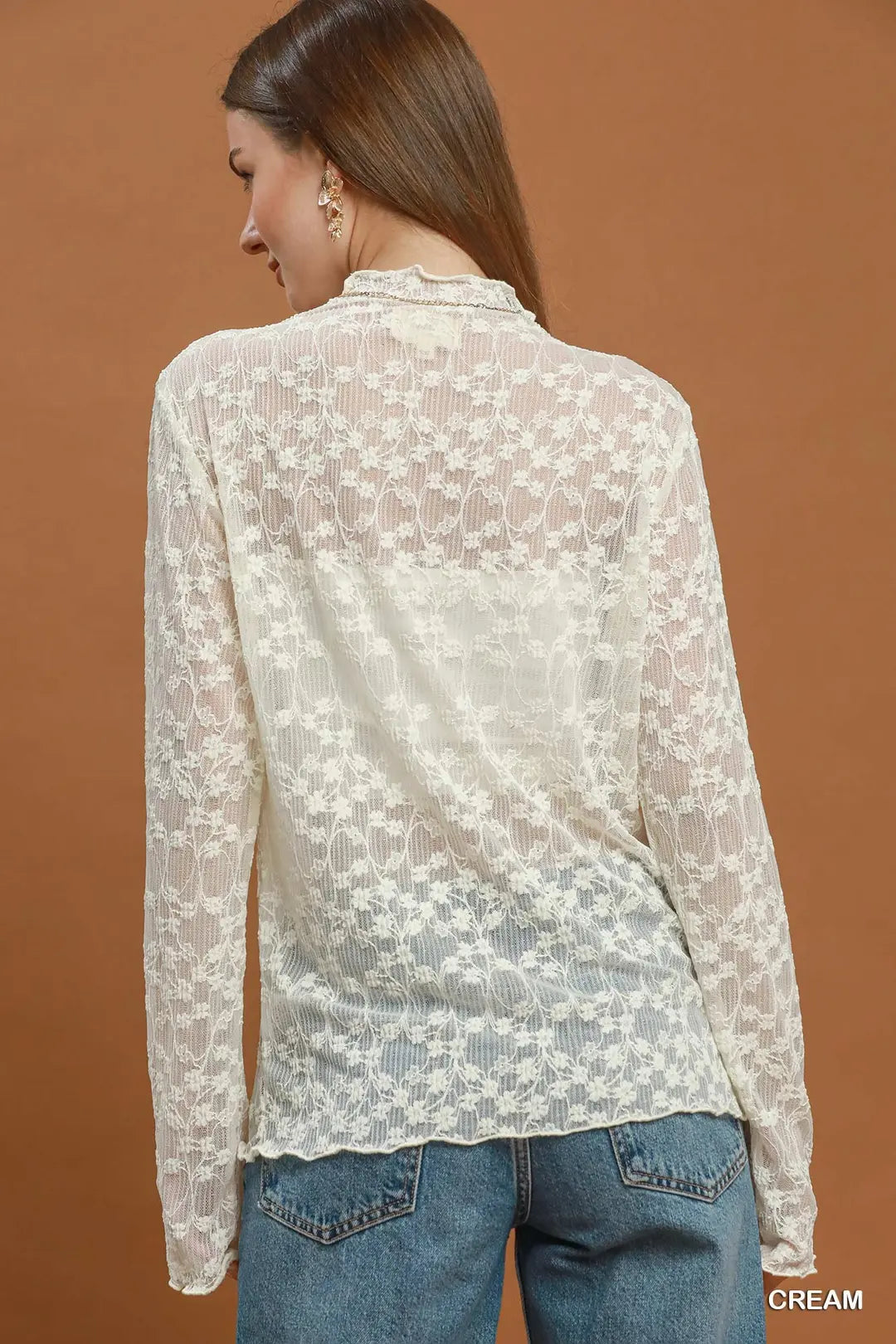 A woman shown from the back wearing a cream lace long-sleeve blouse and blue jeans, standing against a solid warm brown background. The word 'CREAM' appears in the bottom right corner.