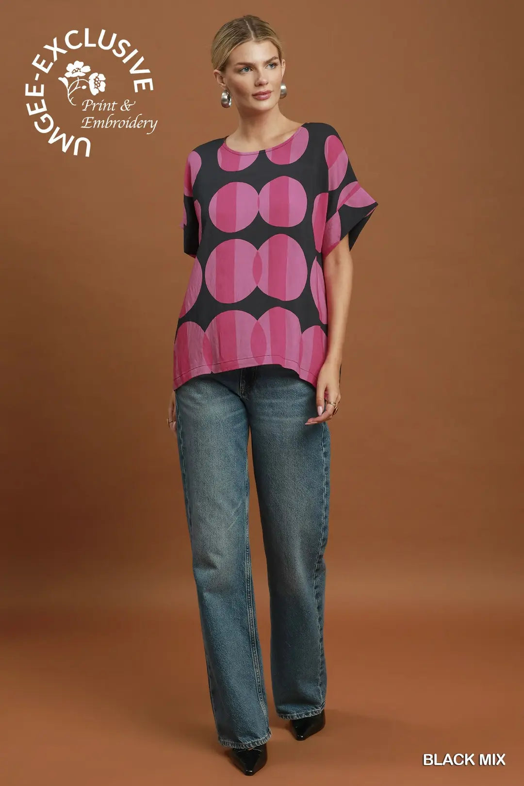 A woman modeling a short-sleeve black top with a bold pink and magenta circular pattern, paired with blue jeans, against a solid brown studio background. A logo for 'Unbee Exclusive Print & Embroidery' is visible in the top left, and 'BLACK MIX' is written in the bottom right.