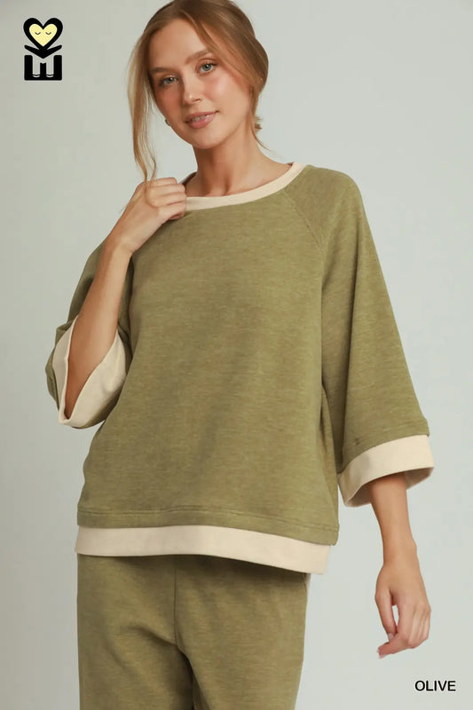 A woman wearing a loose olive green top with contrasting cream trim at the crew neck, sleeve cuffs, and hem, paired with matching olive green pants. The image is set against a solid light gray background with a brand logo in the top-left corner and the word 'OLIVE' in the bottom-right.