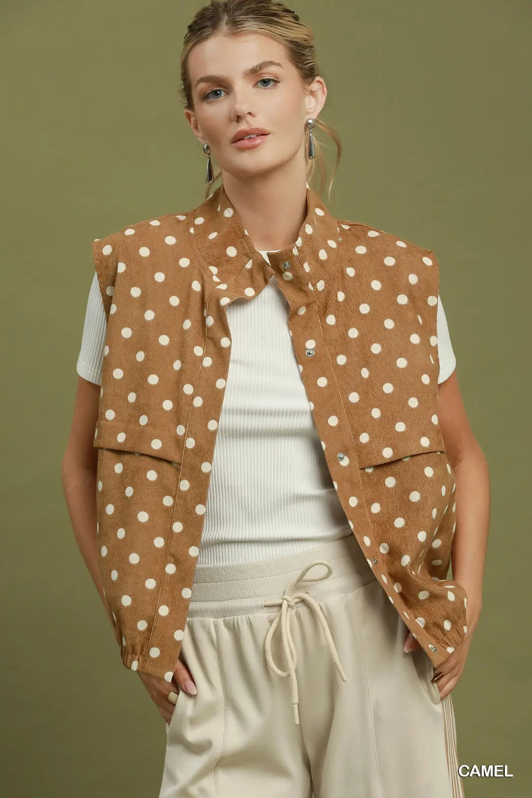 A woman models a sleeveless camel brown vest with white polka dots over a white ribbed top and cream drawstring pants, set against a solid olive green studio background.