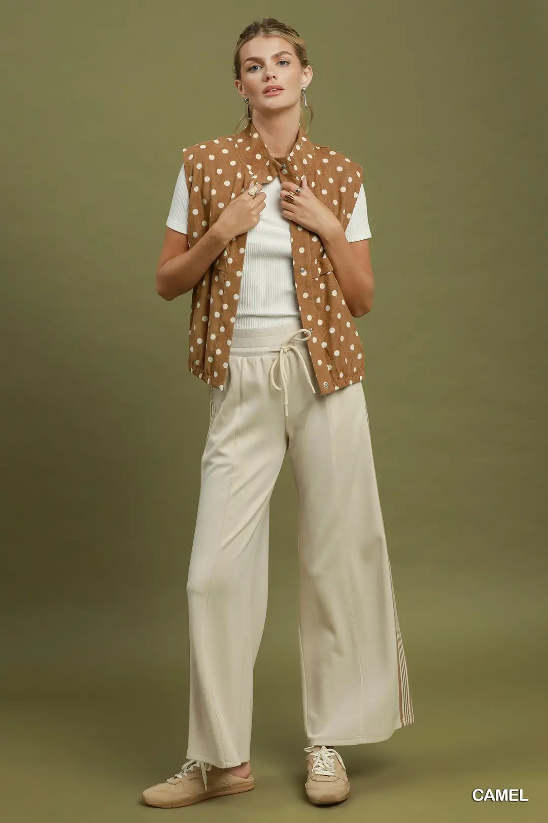A model stands against a solid olive green studio background wearing a camel brown sleeveless vest with white polka dots over a white t-shirt, paired with cream wide-leg trousers and beige sneakers. The word 'CAMEL' is printed in white in the bottom right corner.