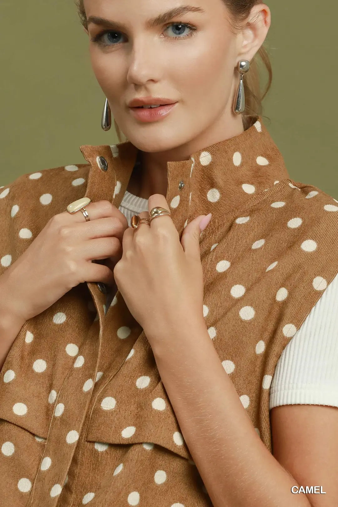 A close-up of a model wearing a camel brown sleeveless vest with white polka dots over a white ribbed top. She is wearing silver dangling earrings and rings, and the background is a solid olive green.