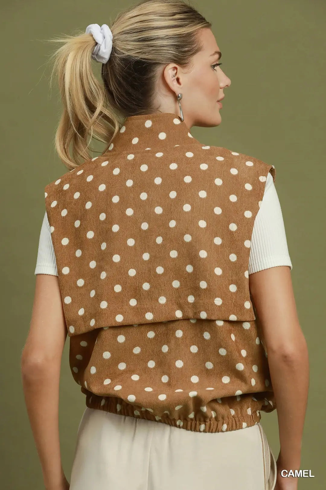 A woman shown from the back wearing a camel brown sleeveless vest with white polka dots over a white ribbed top and cream bottoms, set against a solid olive green background.