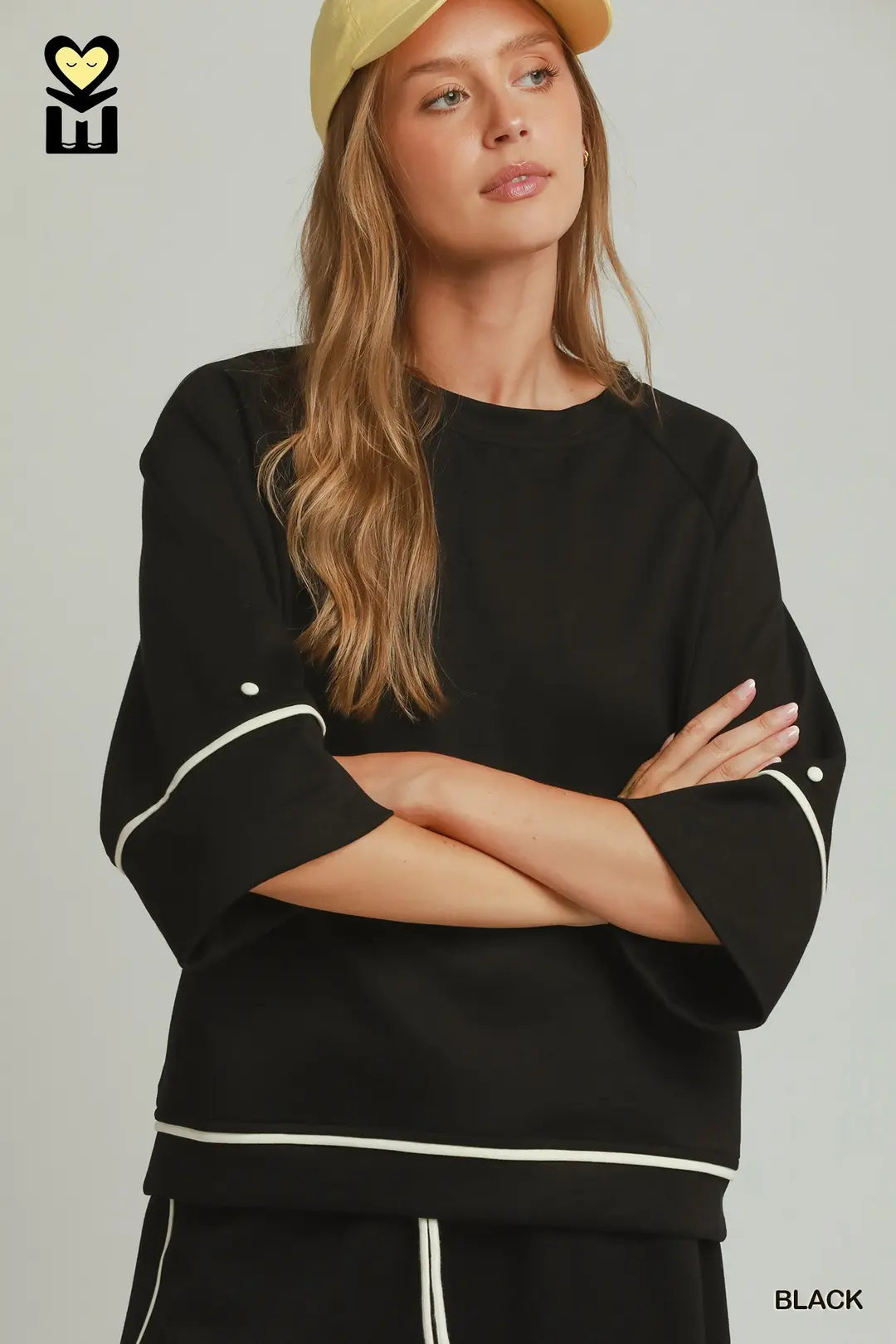 A woman wearing a black crewneck sweatshirt with white contrast piping on the sleeves and hem, paired with black pants and a beige cap, against a light gray studio background.