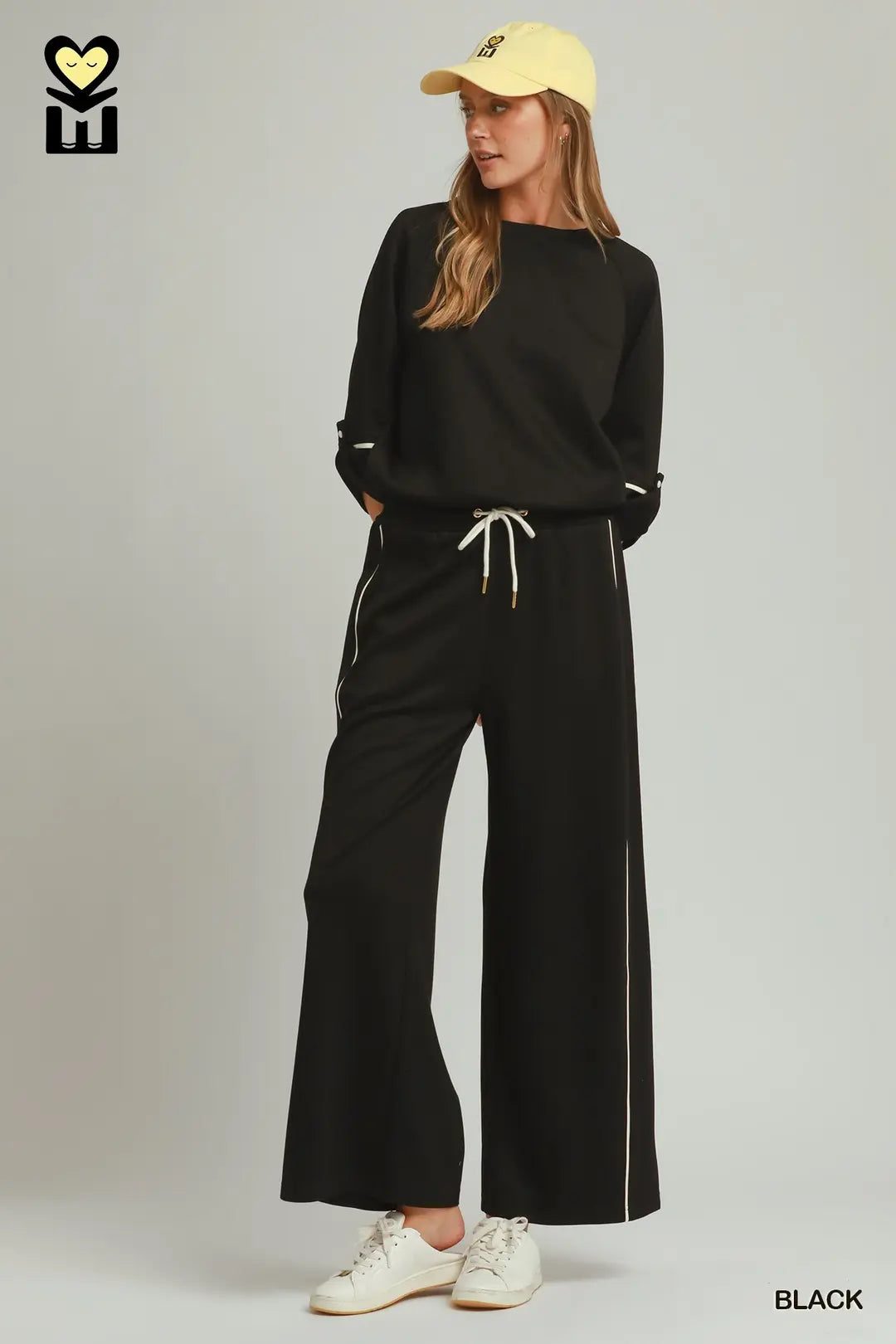A woman modeling a black two-piece lounge set consisting of a long-sleeve sweatshirt and matching wide-leg pants with white drawstrings and piping details, styled with a yellow cap and white sneakers against a light gray studio background.