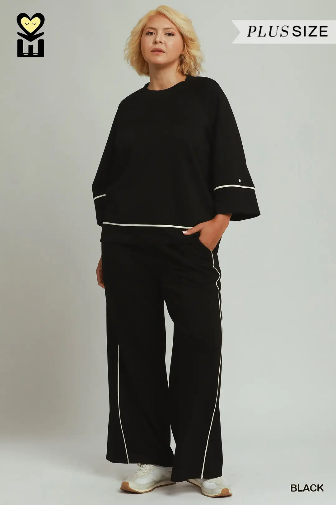 A plus-size model wearing a black two-piece lounge set with white contrast piping details on the sleeves and pants, standing against a light gray studio background.