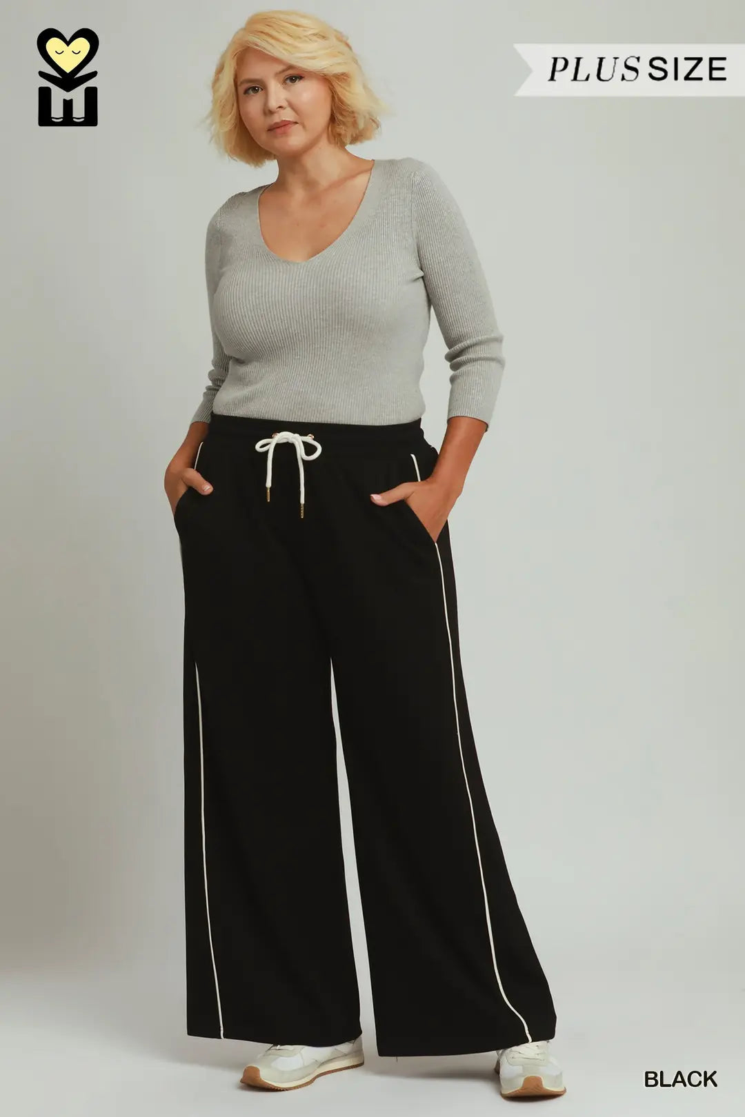A woman modeling black wide-leg sweatpants with white side piping and a white drawstring, paired with a light gray long-sleeve top. The image features a 'PLUS SIZE' banner in the top right and a brand logo in the top left.
