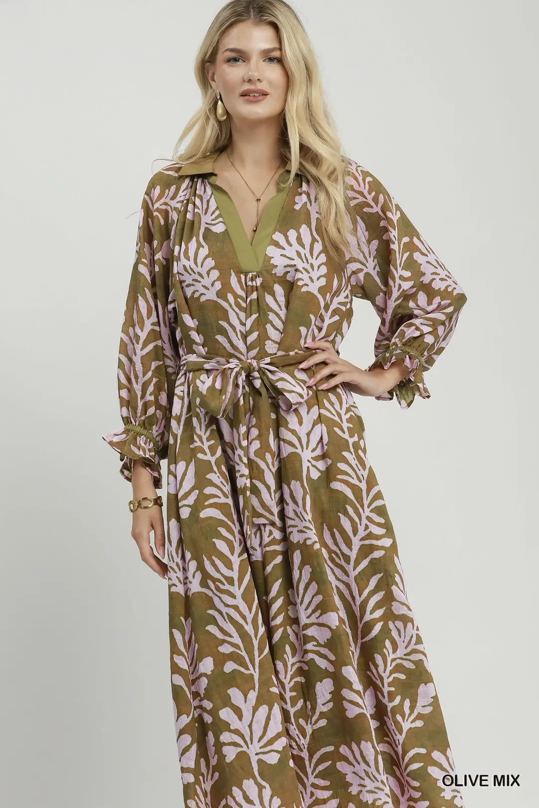 A woman modeling a long-sleeve maxi dress with a tie waist and an olive green and dusty pink botanical leaf pattern, set against a plain light gray studio background.