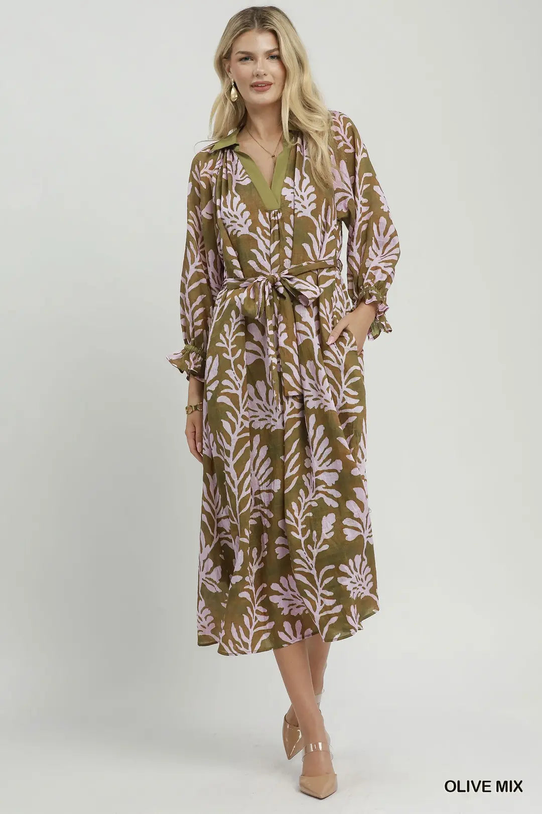 A woman modeling a long-sleeve midi shirt dress with a tie waist, featuring an olive green and dusty pink botanical leaf pattern, set against a plain light gray studio background.