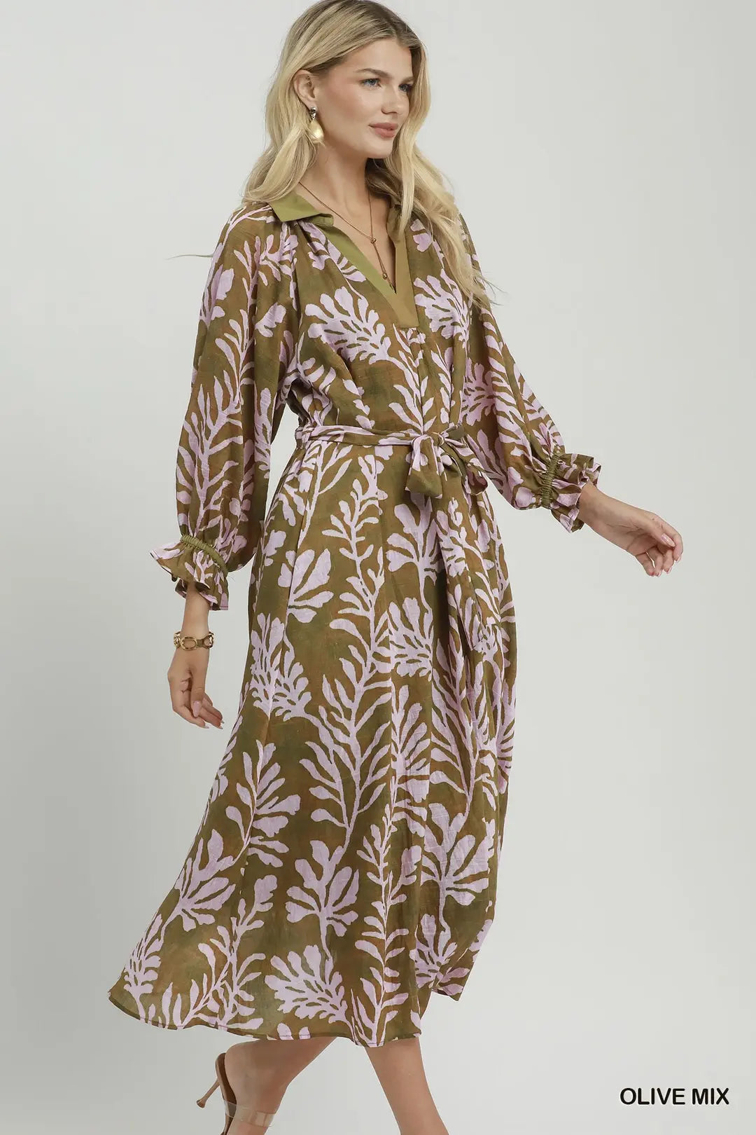 A woman modeling a long-sleeve midi dress with a tie waist and a leafy botanical print in olive green and dusty pink, set against a plain light gray studio background.