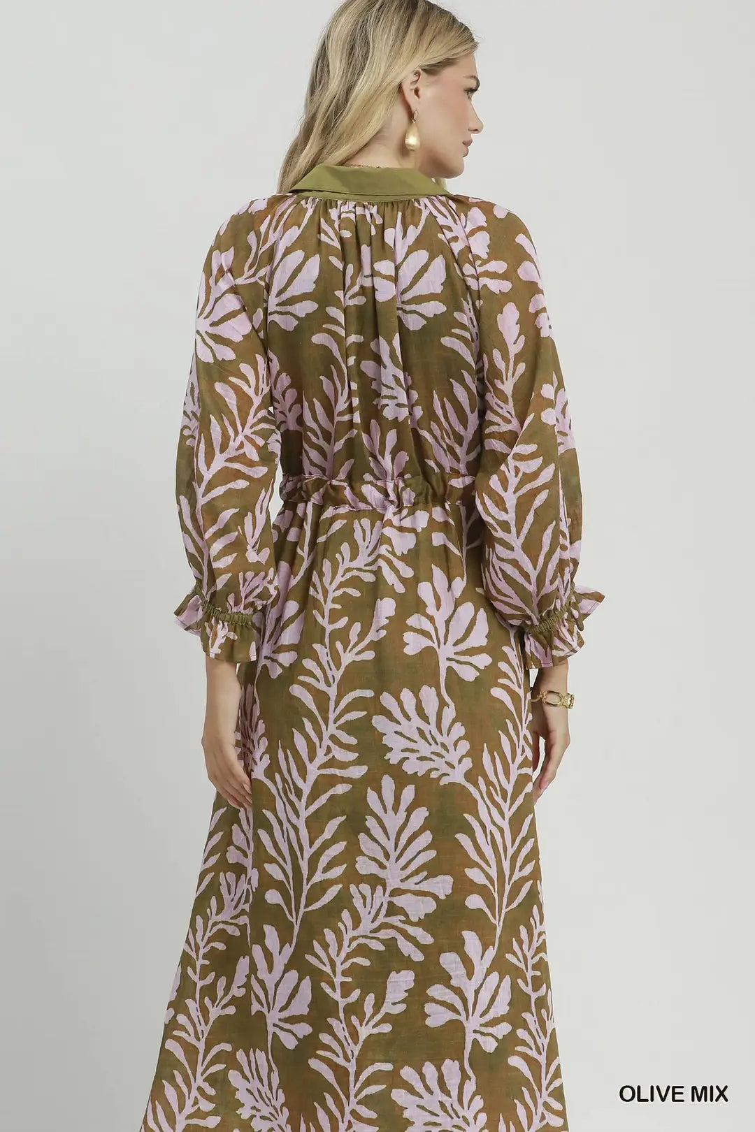 A woman shown from the back wearing a long-sleeve maxi dress with a botanical leaf pattern in olive green and light pink. The dress features a gathered waist and tie detail at the back collar.