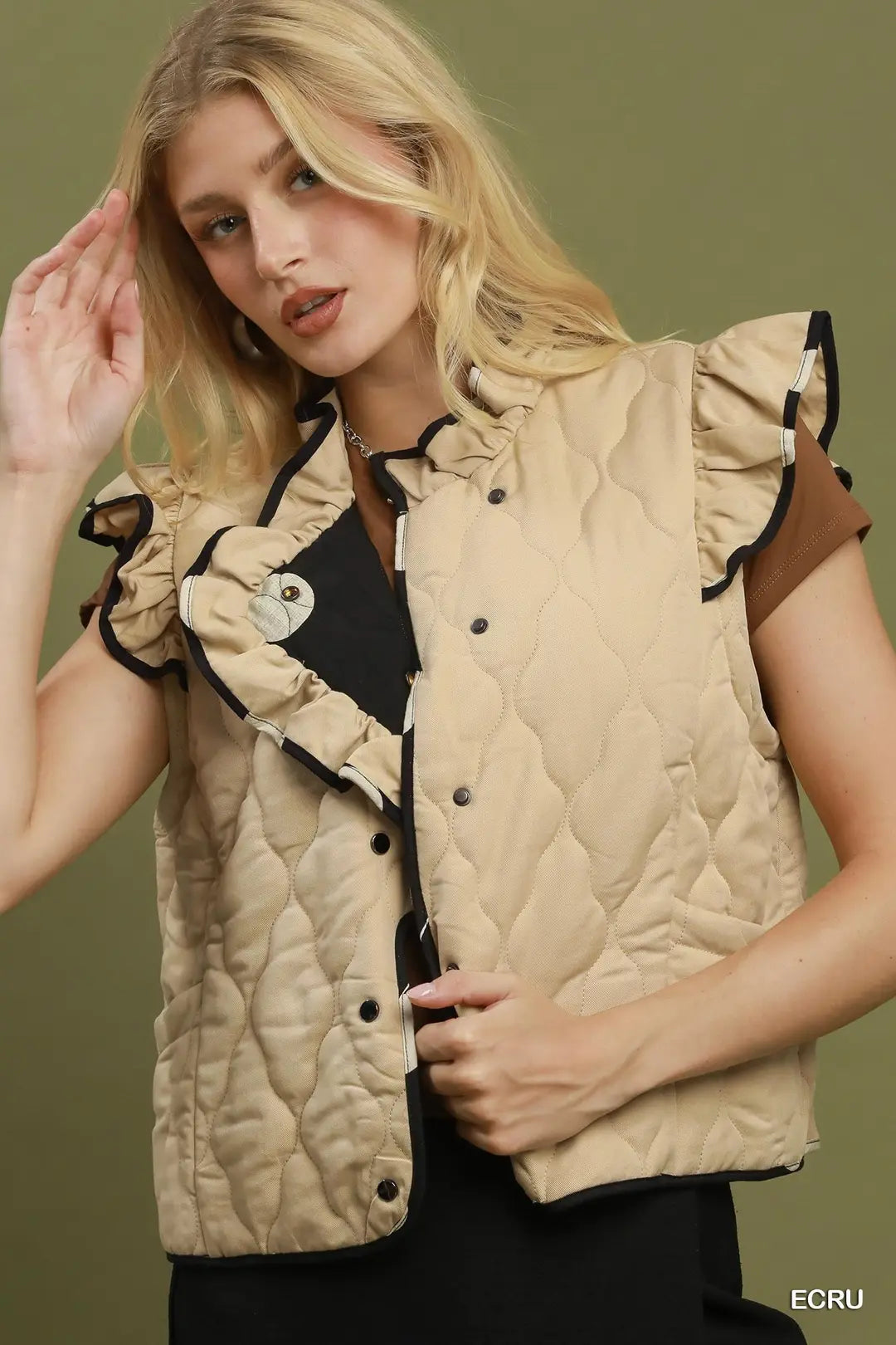 A blonde model wearing a beige quilted sleeveless vest with black trim, ruffled shoulders, and a snap-button front, styled over a black top against an olive green background.