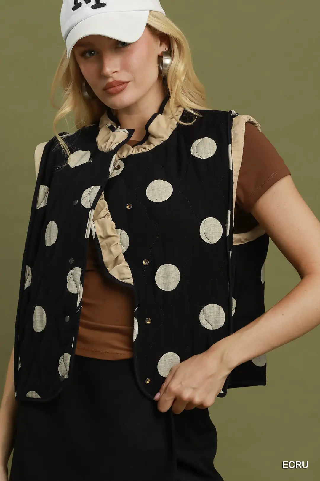 A model wearing a black sleeveless quilted vest with beige ruffle trim and beige polka dots, styled over a brown top and black pants, against an olive green background.