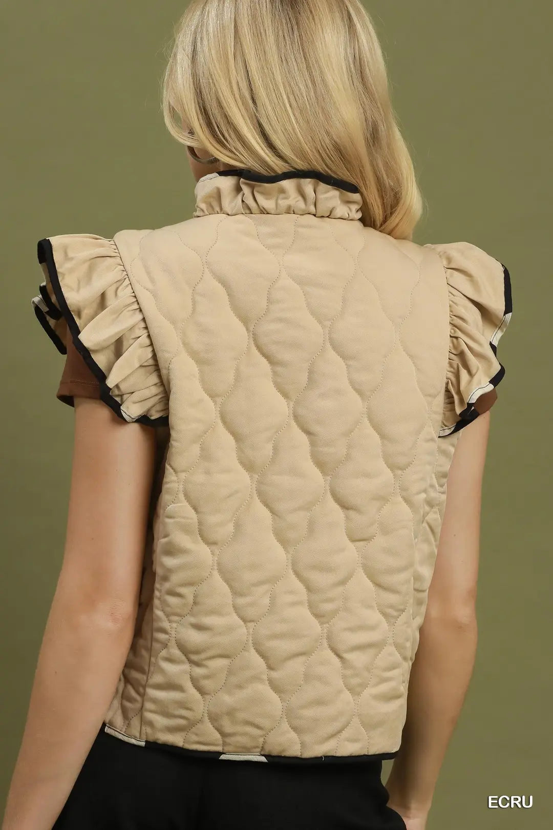 A woman viewed from the back wearing a beige quilted sleeveless vest with ruffled cap sleeves and black trim, against an olive green background.
