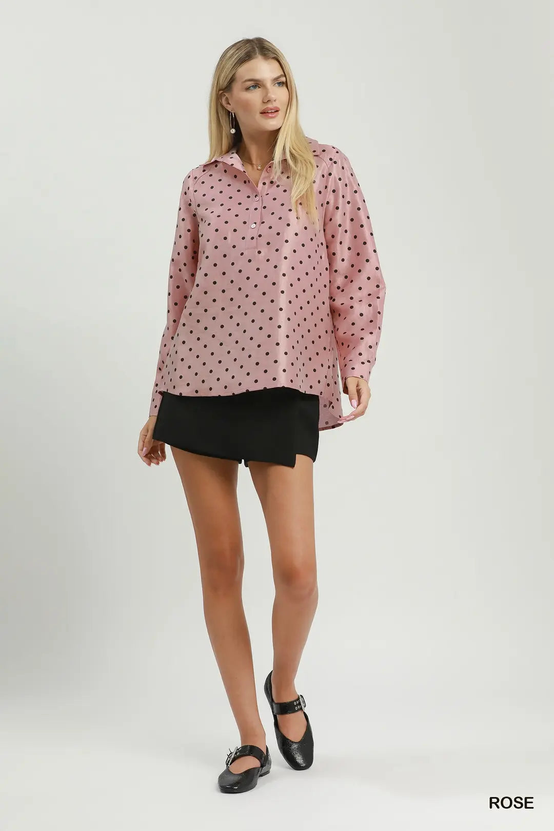 A woman modeling a pink long-sleeve button-up shirt with a black polka dot pattern, paired with a black wrap-style mini skirt and black flat shoes, against a light gray studio background.