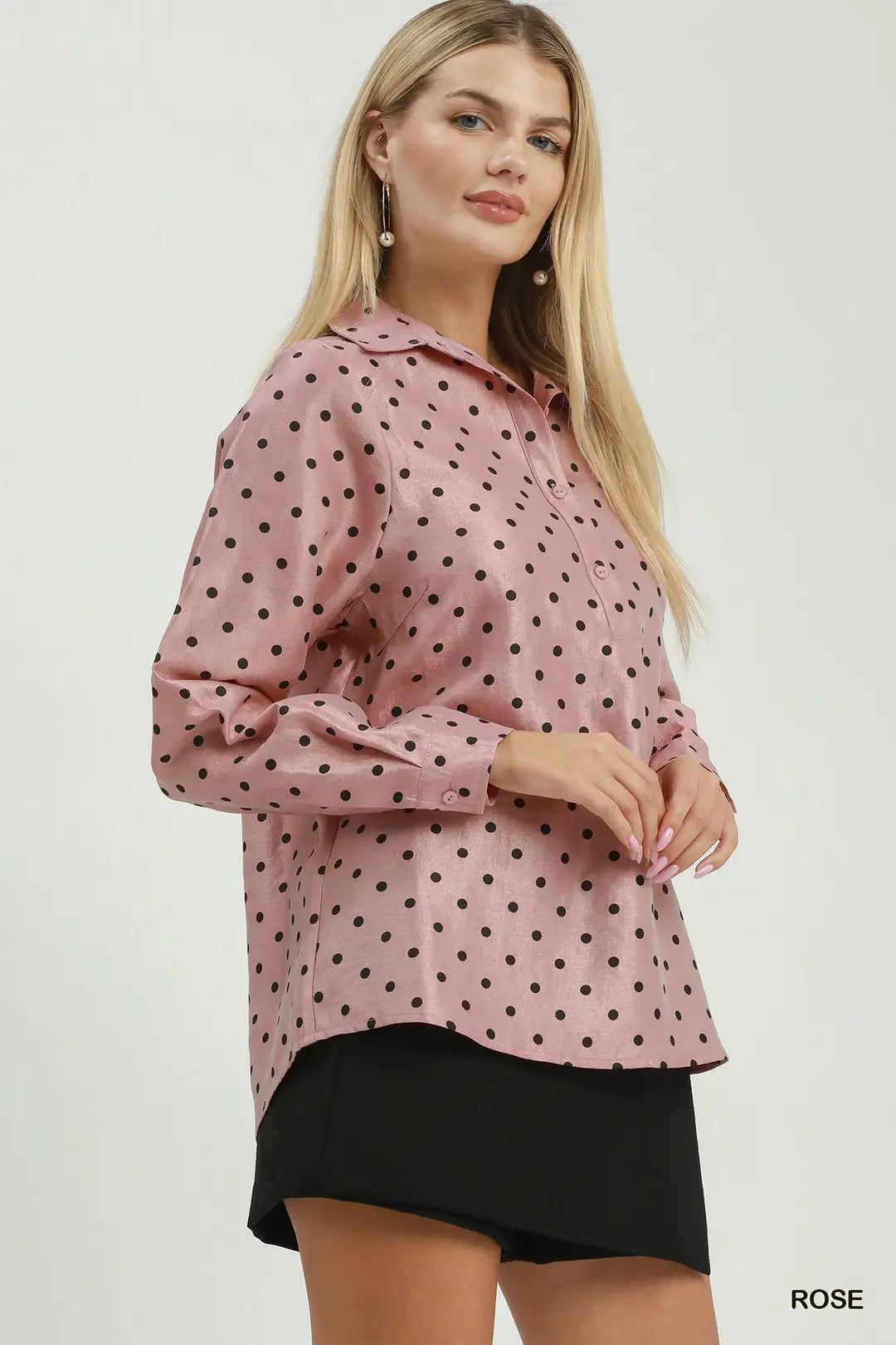 A woman modeling a pink long-sleeve button-up shirt with black polka dots, paired with black shorts, against a plain light gray studio background.