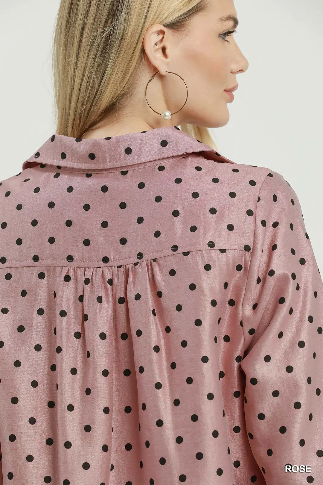 A woman shown from the back wearing a dusty pink long-sleeve blouse with black polka dots. She is also wearing large gold hoop earrings.