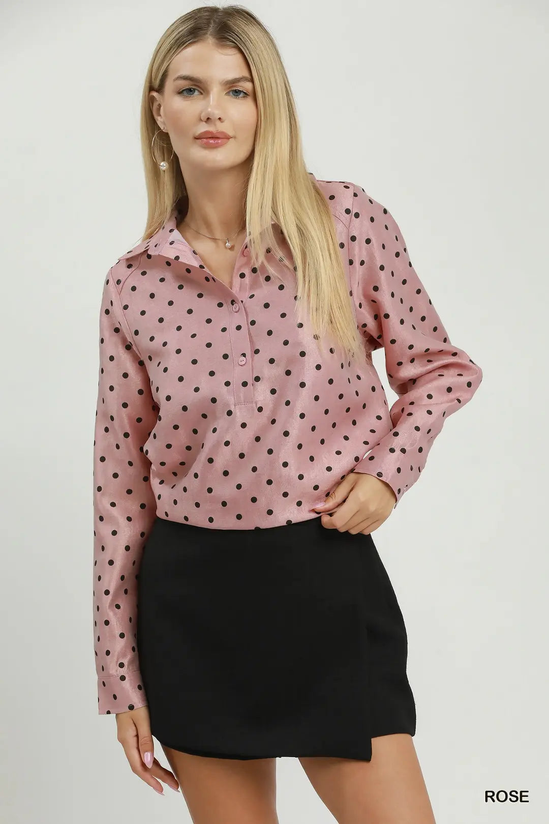 A woman modeling a pink long-sleeve button-up blouse with black polka dots, paired with a black mini skirt, against a plain light gray studio background.