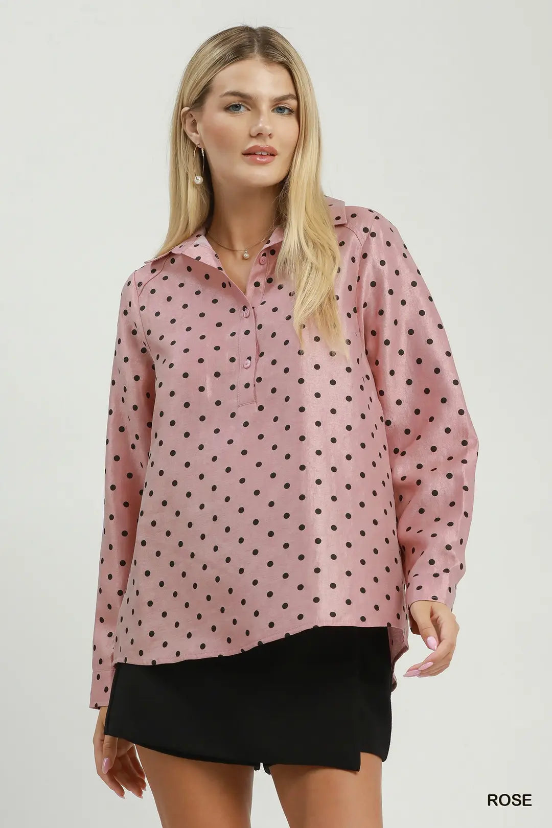 A woman modeling a long-sleeve collared blouse in dusty rose pink with a black polka dot pattern, paired with black shorts, against a light gray studio background.