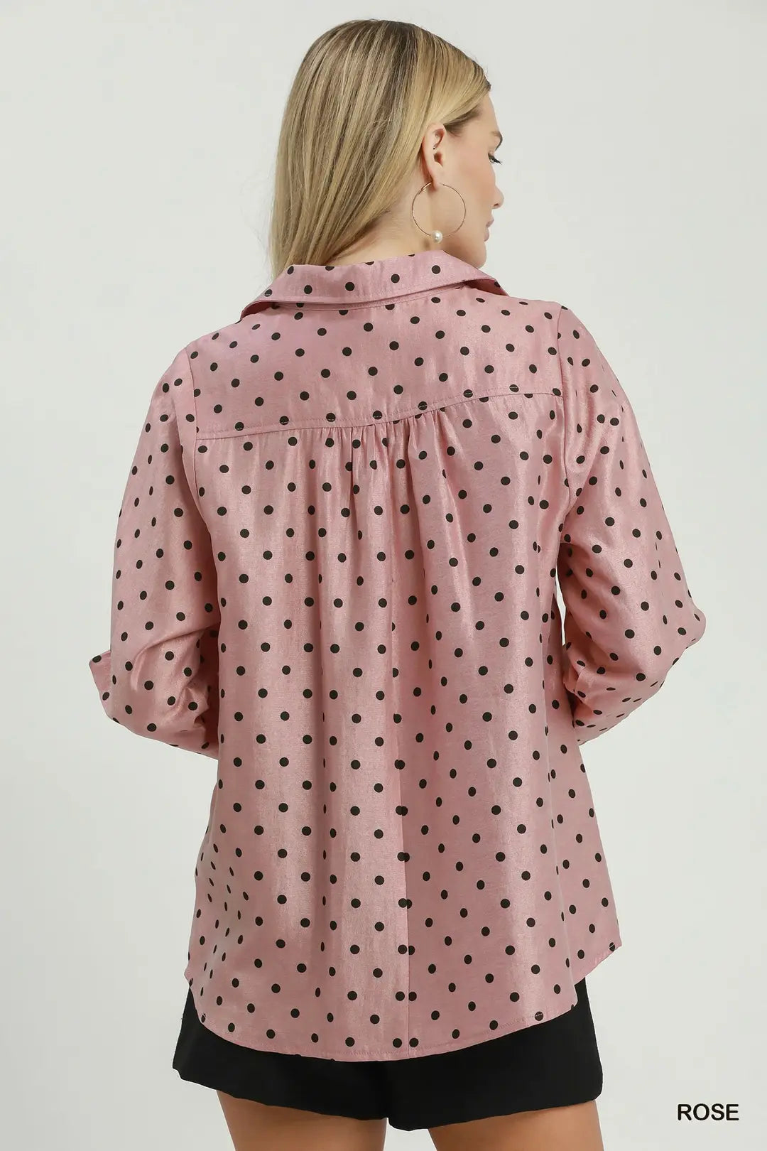 A woman shown from the back wearing a pink button-up shirt with black polka dots, paired with black shorts, against a plain light gray studio background.