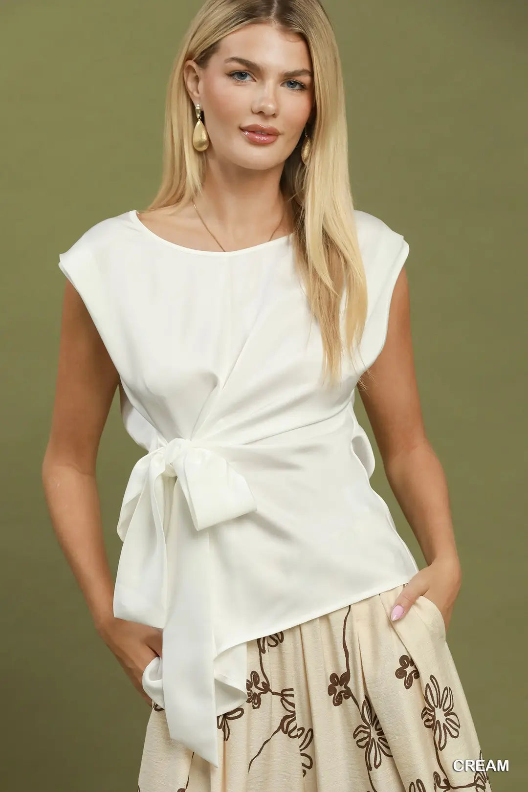 A woman modeling a white sleeveless wrap-style top with a tied waist bow, paired with a cream floral skirt, against a solid olive green background.