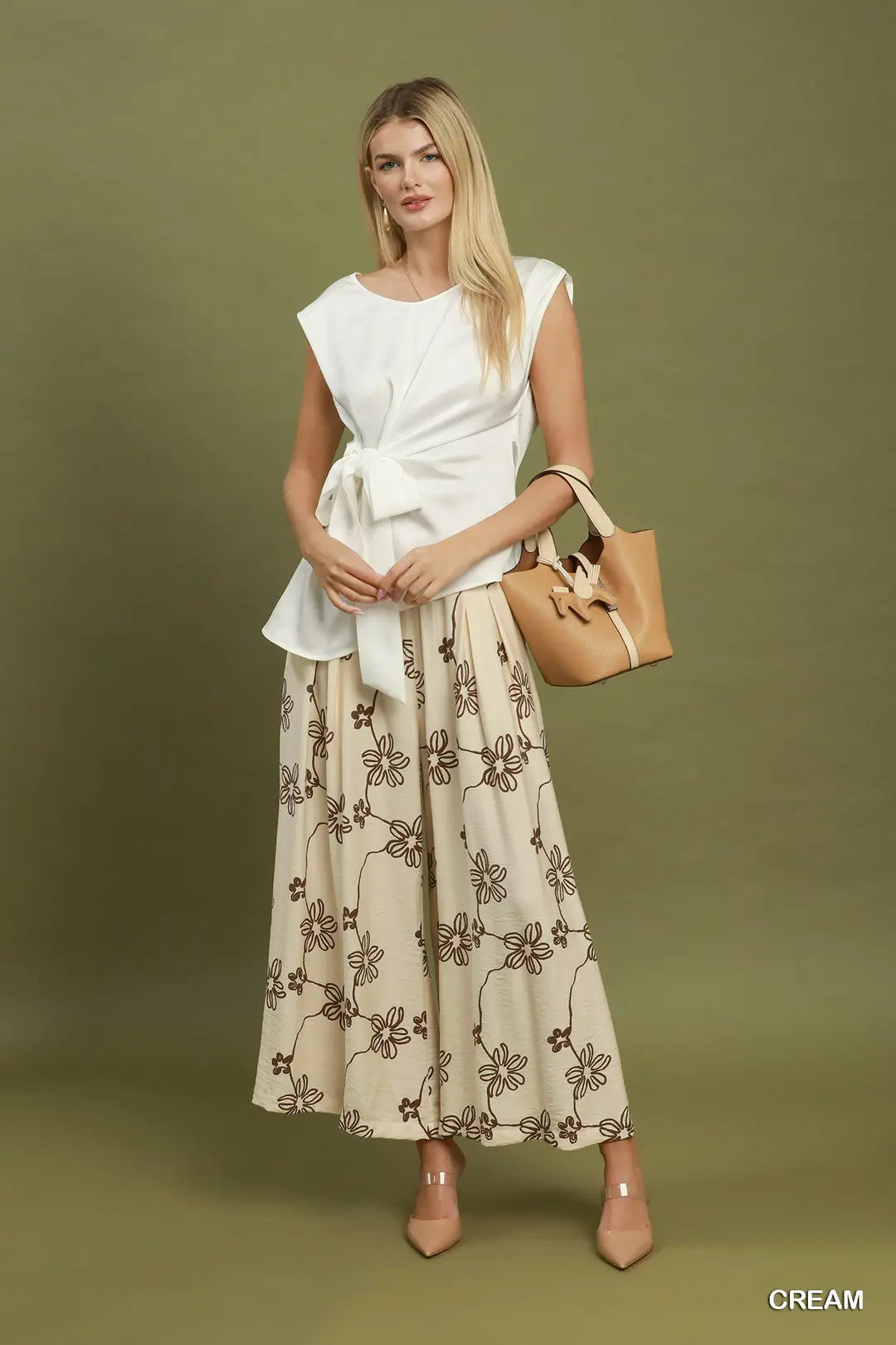 A woman models a two-piece outfit consisting of a white sleeveless wrap-style top and a cream maxi skirt with a brown floral pattern, holding a tan handbag against a solid olive green studio background.