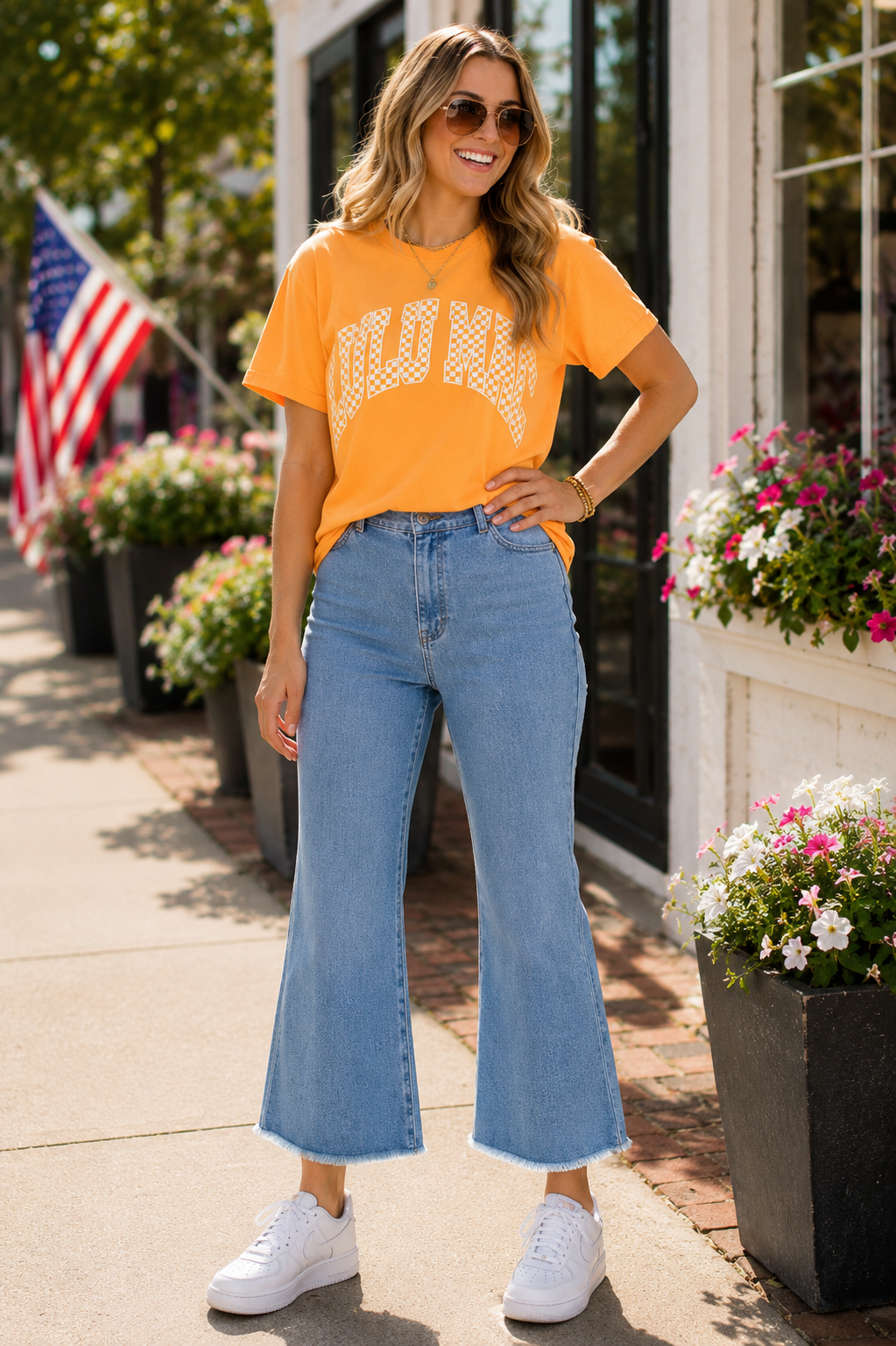 A woman stands outdoors on a sidewalk wearing a bright orange graphic T-shirt with white lettering, light blue high-waisted flare jeans, and white sneakers. Flower boxes and an American flag are visible in the background.