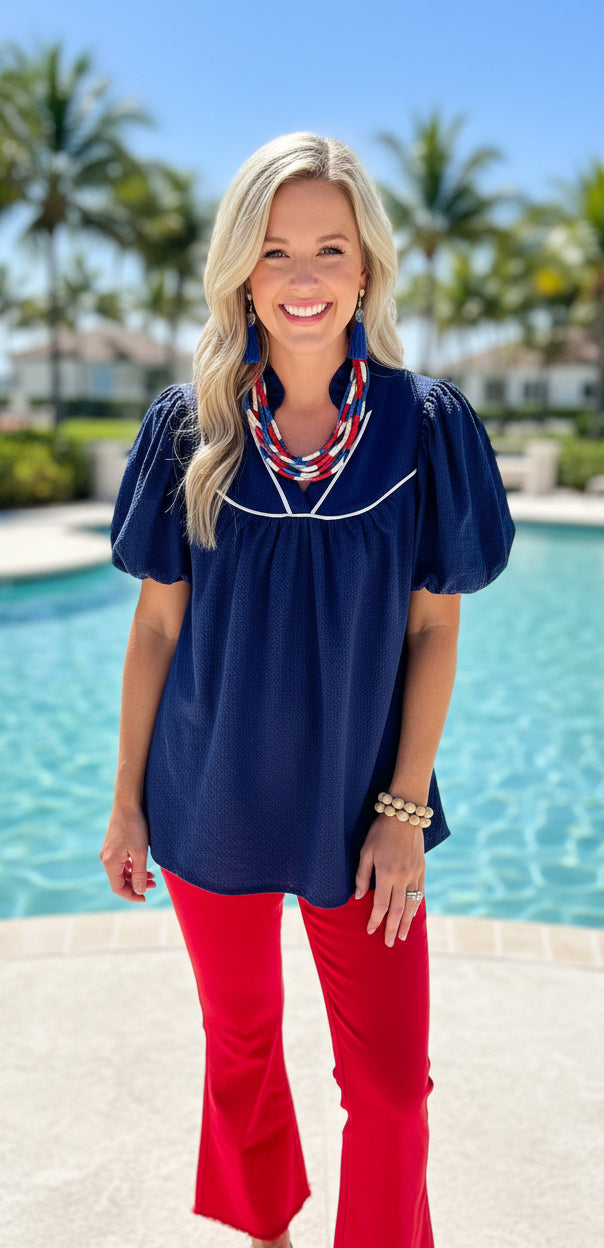 Woman in a boutique navy top and red pants standing by a pool with palm trees in the background