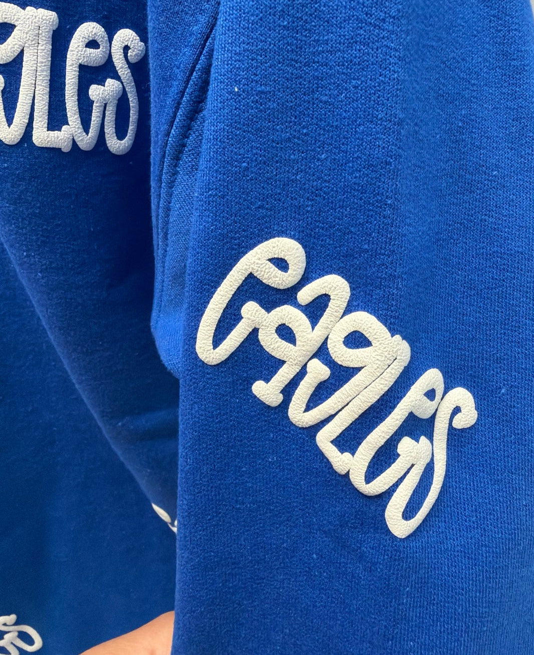 Close-up of a royal blue sweatshirt sleeve featuring a large white embroidered script logo that reads 'Eagles' on the upper arm, with a hand holding the sleeve at the bottom.