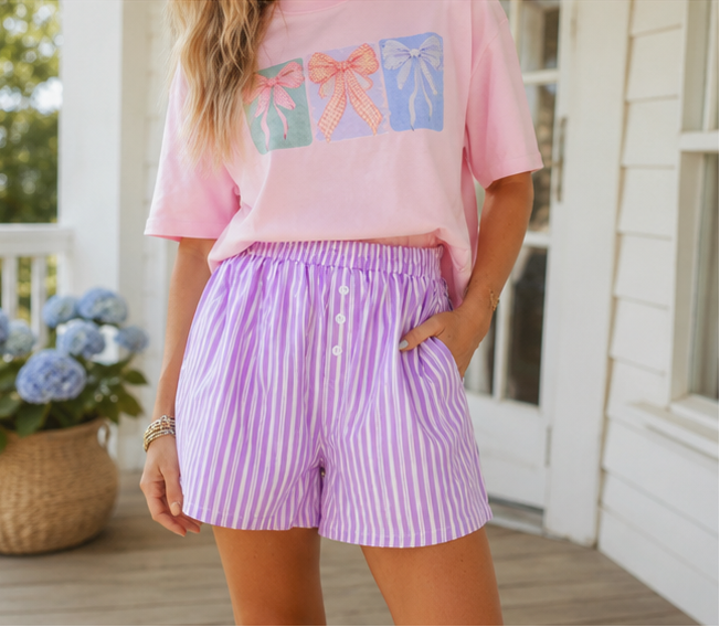 Person wearing a pink shirt with bow designs and purple striped shorts on a porch.