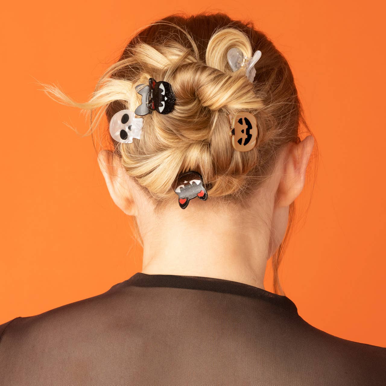 A person shown from the back with blonde hair in a messy bun, wearing several small Halloween-themed hair clips against a solid orange background.