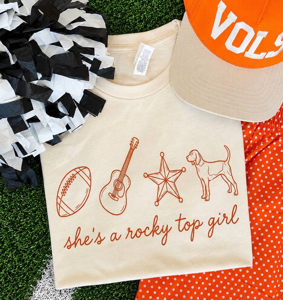 A cream-colored graphic t-shirt featuring orange line drawings of a football, guitar, star, and dog, with the text 'she's a rocky top girl' in orange script. The shirt is styled in a flat-lay arrangement on artificial green turf alongside a black and white pom-pom, an orange and white polka dot shirt, and an orange and white cap.