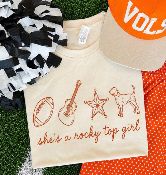A cream-colored graphic t-shirt featuring orange line drawings of a football, guitar, star, and dog, with the text 'she's a rocky top girl' in orange script. The shirt is styled in a flat-lay arrangement on artificial green turf alongside a black and white pom-pom, an orange and white polka dot shirt, and an orange and white cap.