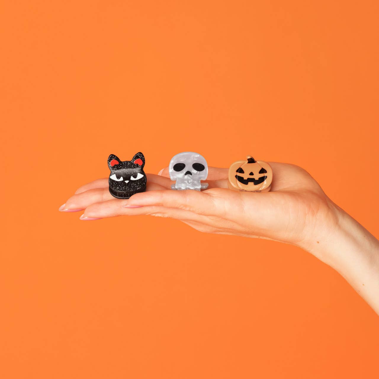 Three small Halloween-themed figurines, including a black cat, a white skull, and a jack-o'-lantern, displayed on a person's open hand against a solid orange background.