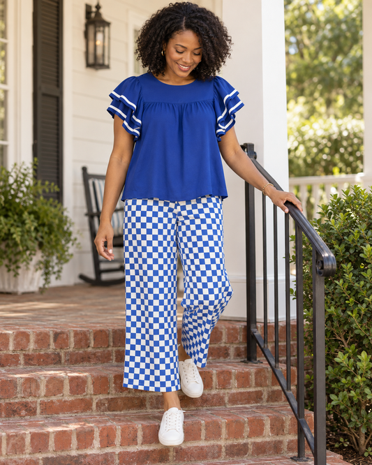 A woman walking down brick porch steps wearing a royal blue ruffle-sleeve top and blue-and-white checkered wide-leg pants, with white sneakers. The photo is taken outdoors on a home porch with plants and a railing visible.
