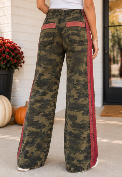 A person wearing wide-leg camouflage pants with red side stripes and red back pocket accents, standing outdoors near a white brick wall and pumpkins.