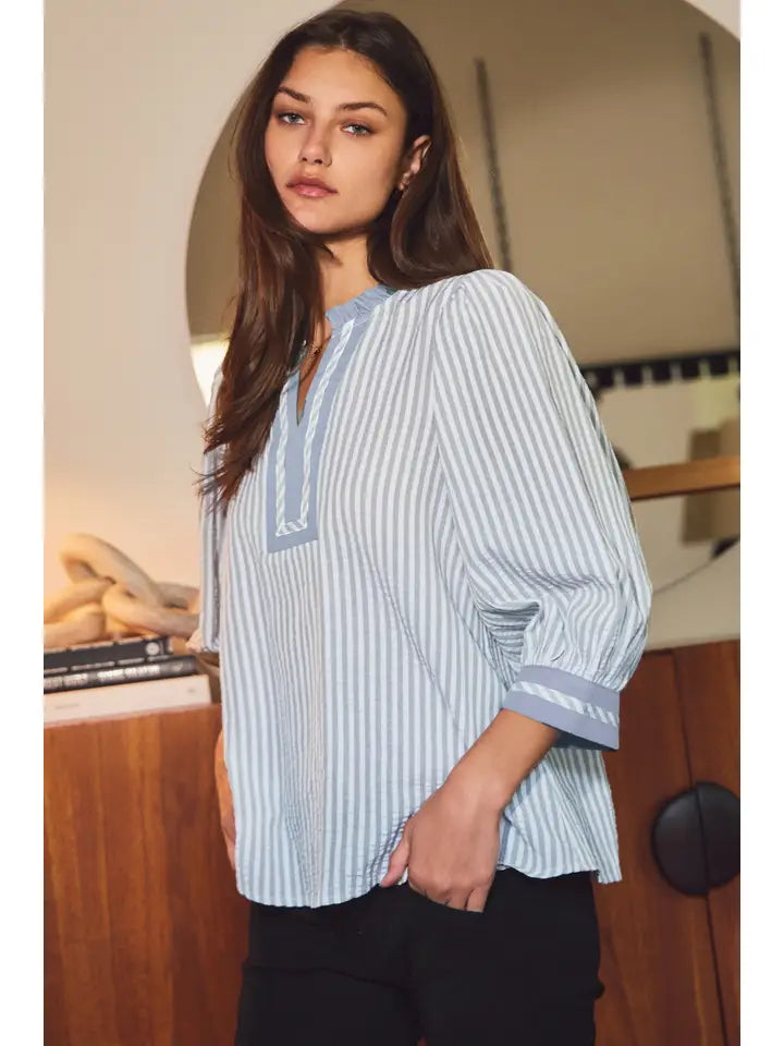 A woman wearing a light blue and white vertically striped blouse with a V-neckline and three-quarter sleeves, paired with black pants, in an indoor setting.