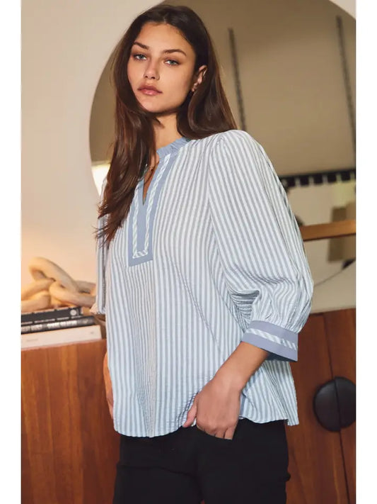 A woman wearing a light blue and white vertically striped blouse with a V-neckline and three-quarter sleeves, paired with black pants, in an indoor setting.