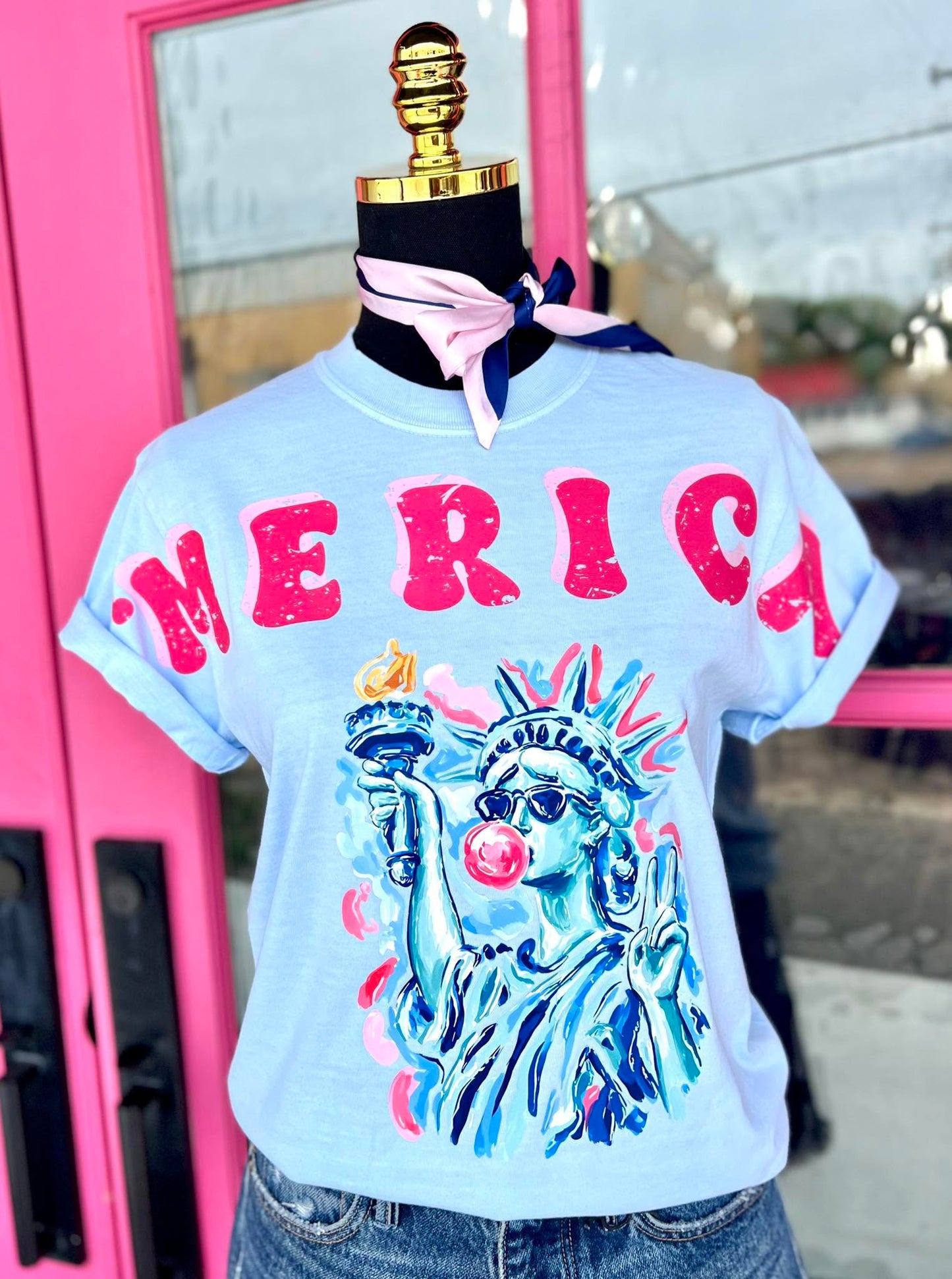 Light blue t-shirt with 'MERICA' and Statue of Liberty graphic on a mannequin against a pink door.