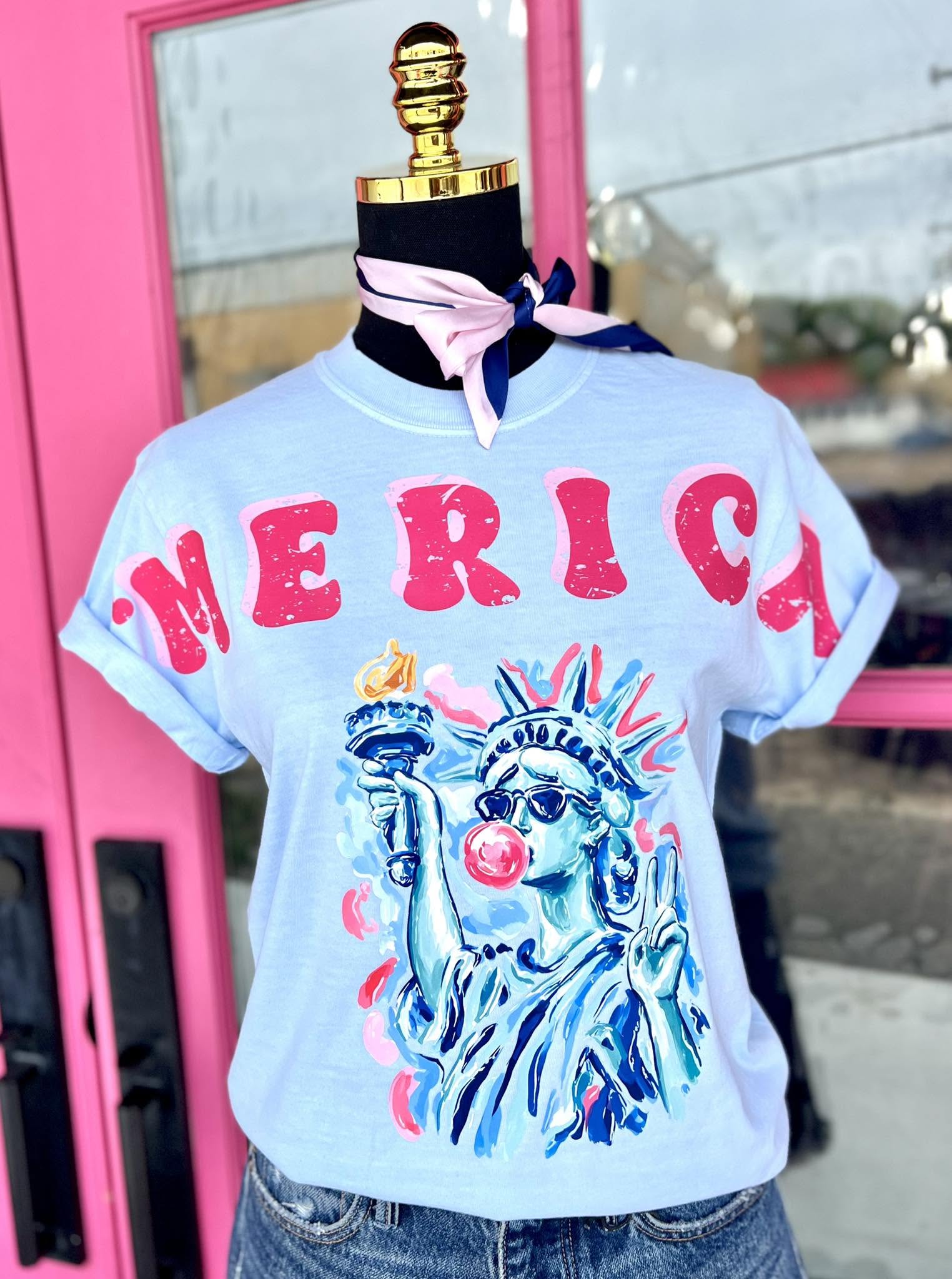 Light blue t-shirt with 'MERICA' and Statue of Liberty graphic on a mannequin against a pink door.
